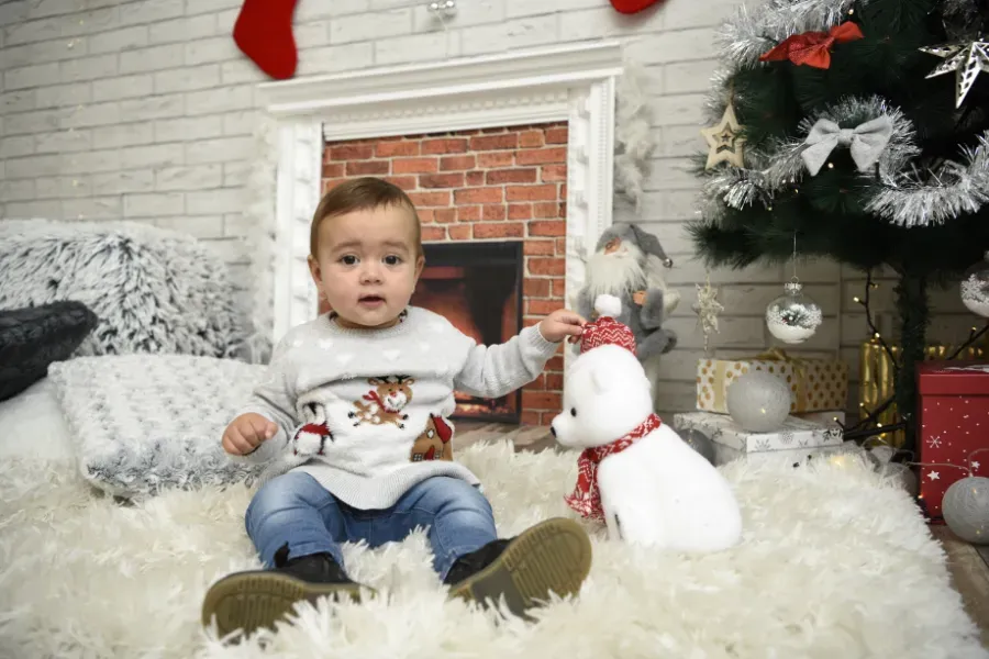 Un bebé sentado sobre una alfombra mullida junto a un muñeco de nieve y un árbol de Navidad en una acogedora habitación navideña.