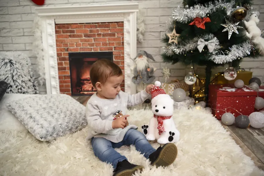 Un niño sentado en una alfombra mullida junto a un árbol de Navidad, intentando alcanzar un pequeño muñeco de nieve de juguete junto a una chimenea.
