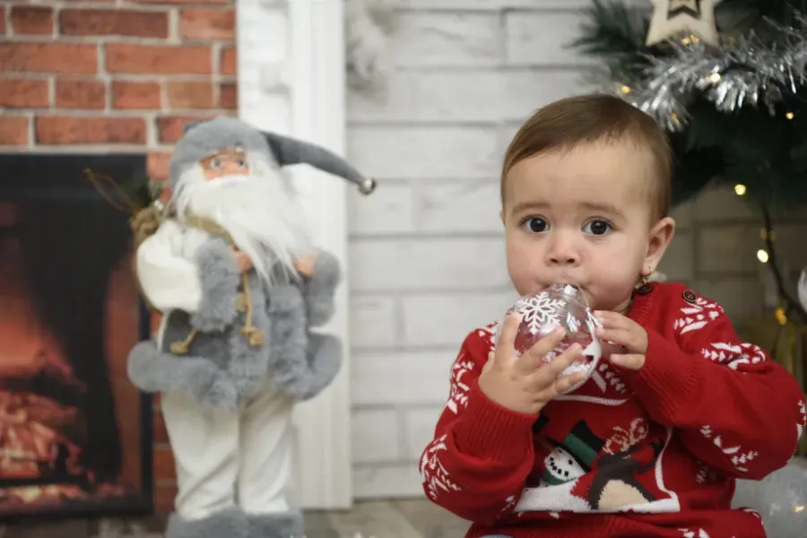 Bebé con suéter rojo navideño sosteniendo un adorno brillante junto a una decoración de Papá Noel y un árbol de Navidad.