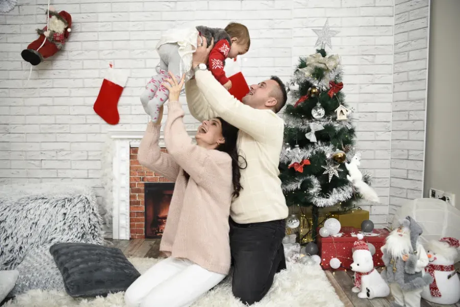 Un adulto y un niño jugando con un bebé junto a un árbol de Navidad en una acogedora sala de estar navideña.