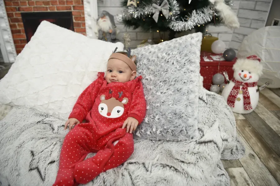 Un bebé con un pelele rojo navideño yace en una cama con almohadas blancas, junto a un árbol de Navidad y una decoración de muñeco de nieve.