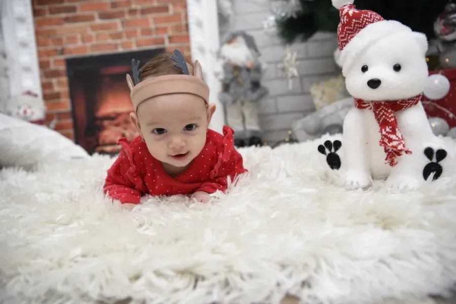 Un bebé con un conjunto rojo yace sobre una alfombra blanca y mullida junto a un osito de peluche blanco en un acogedor ambiente navideño.