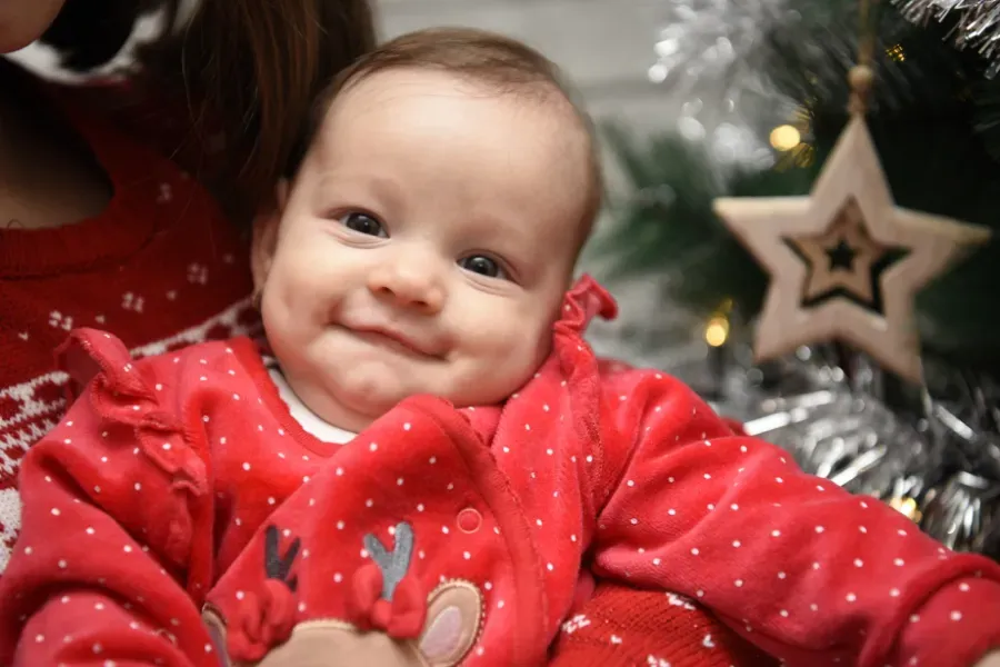 Bebé sonriente con un suéter rojo navideño y un árbol de Navidad y un adorno de estrella al fondo.