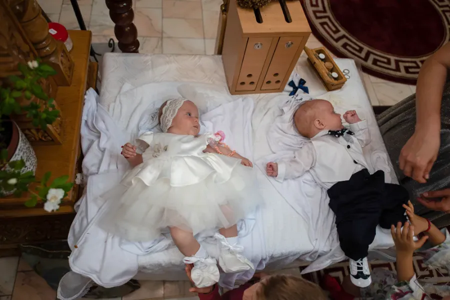 Dos bebés acostados sobre una manta blanca, uno con un vestido blanco y el otro con pantalones negros, con las manos cerca
