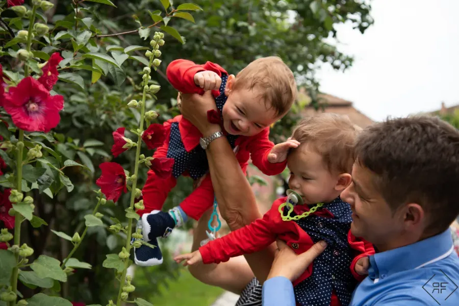Dos niños son alzados para recoger flores rojas en un jardín verde, con un adulto ayudándoles cerca.