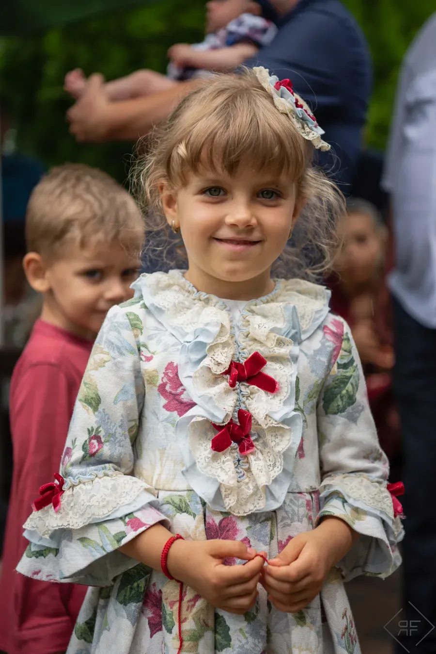 Niña con un vestido floral con detalles de cinta roja, de pie al aire libre con otros niños borrosos detrás de ella.