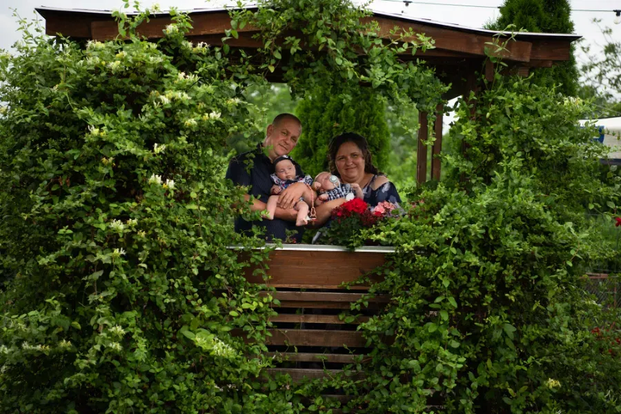 Familia asomándose por una ventana enmarcada por un denso follaje verde, con rostros sonrientes y plantas en macetas cerca.