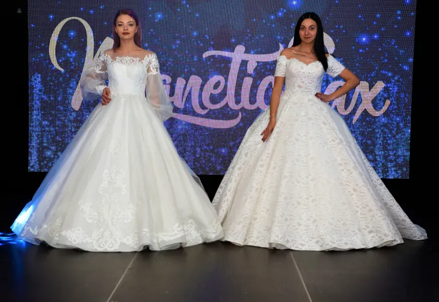 Dos mujeres con vestidos de gala blancos posan en un escenario con un telón de fondo azul estrellado y un letrero del evento en letra cursiva.
