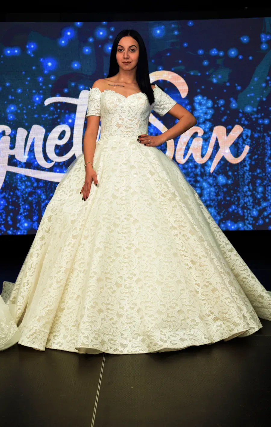 Mujer con un vestido de gala blanco de pie sobre un escenario con un fondo azul brillante.