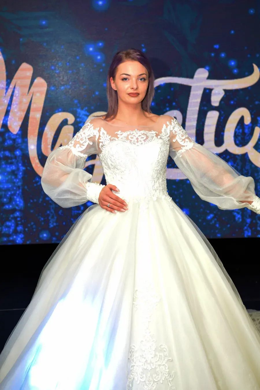 Mujer con un vestido de gala blanco posando en una pasarela con un fondo iluminado en azul.