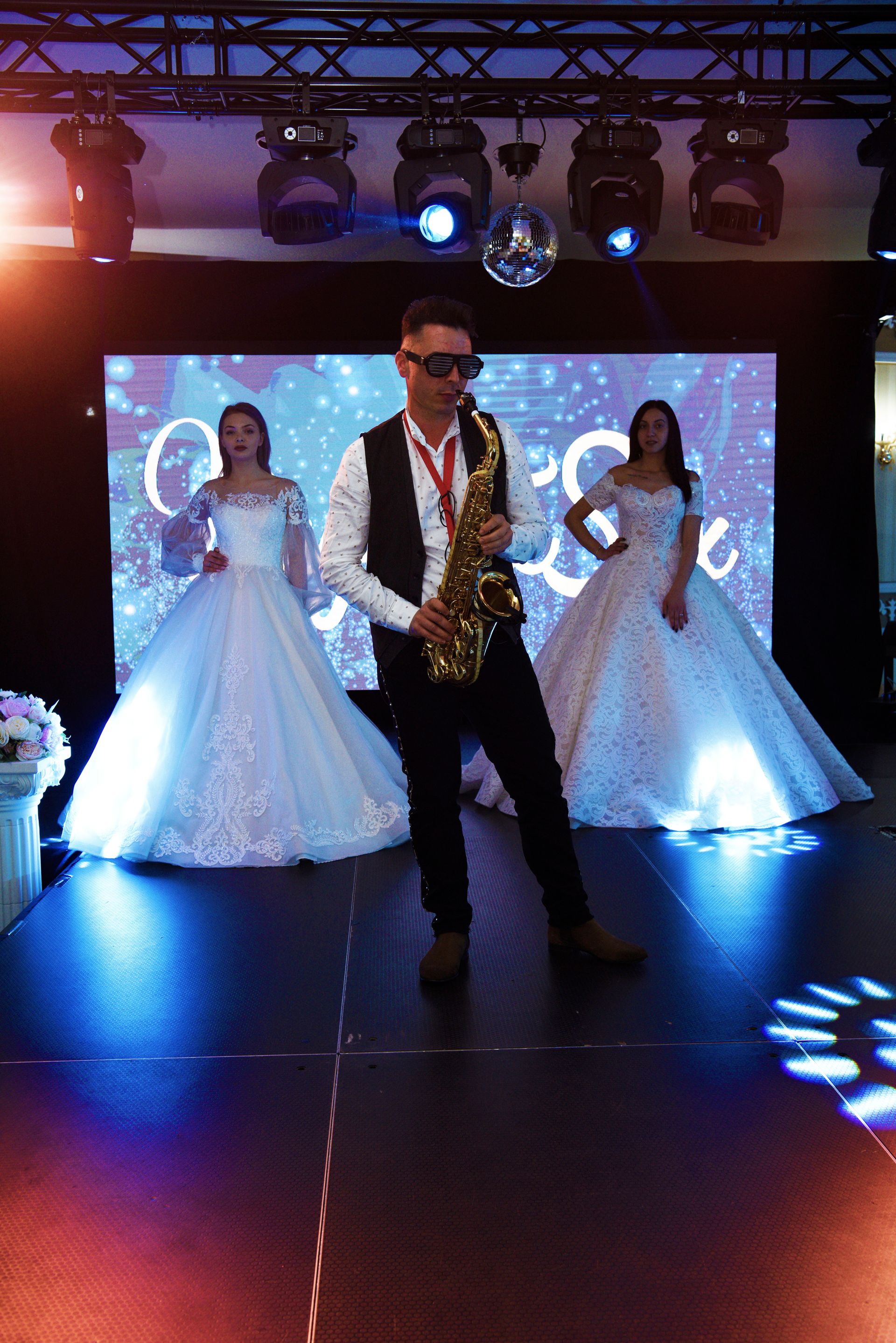 Un cantante con gafas de sol actuando en el escenario con dos novias vestidas de blanco detrás de él.