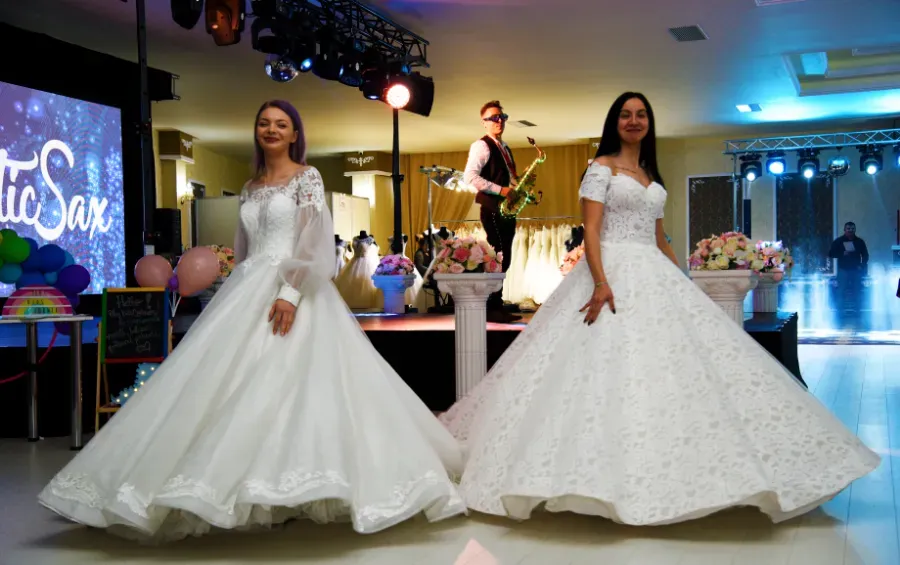 Dos mujeres con vestidos de gala blancos posan en un escenario durante un evento formal, con un músico actuando detrás de ellas.