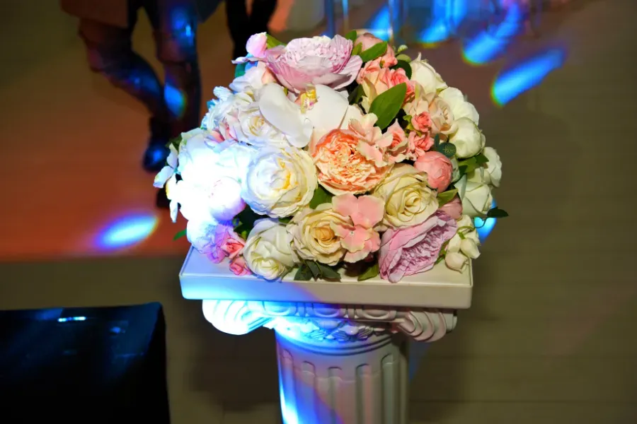 Un ramo de flores en tonos pastel sobre un pedestal blanco, iluminado por luces de colores azul y rosa.
