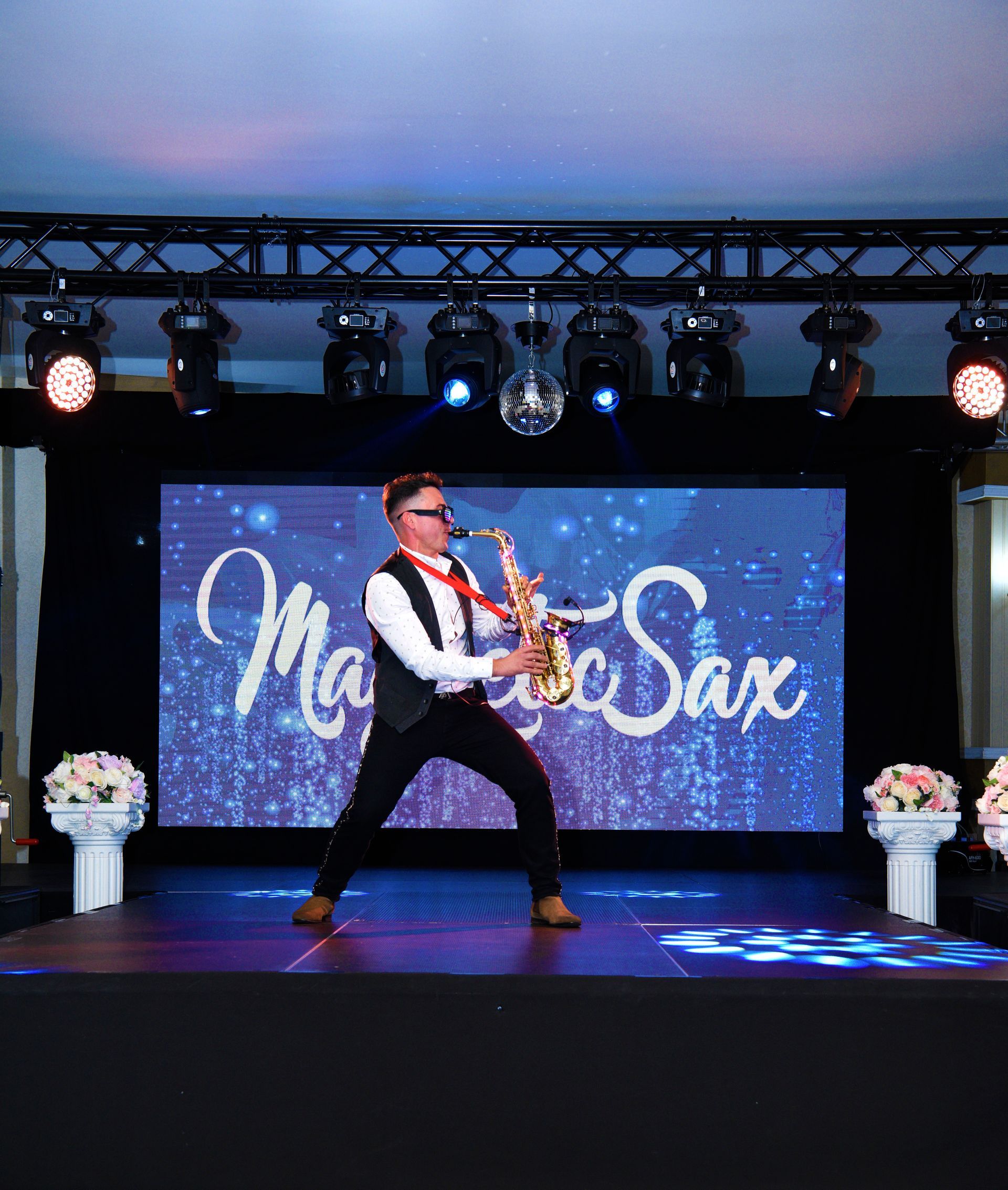 Músico tocando el saxofón en un escenario iluminado con un telón de fondo de “Merry Sax” y decoraciones florales.