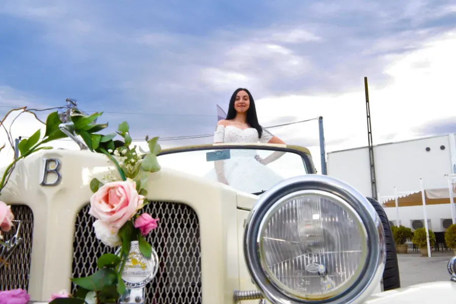 Novia de pie en un coche antiguo blanco decorado con flores rosas bajo un cielo nublado.