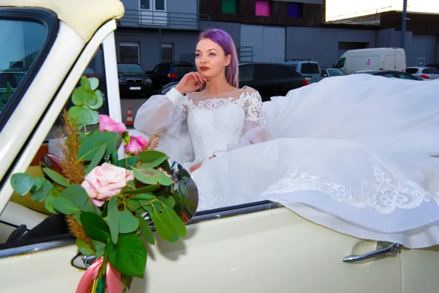 Novia con vestido blanco recostada sobre un coche color crema, sosteniendo un teléfono, con un ramo de flores en primer plano.