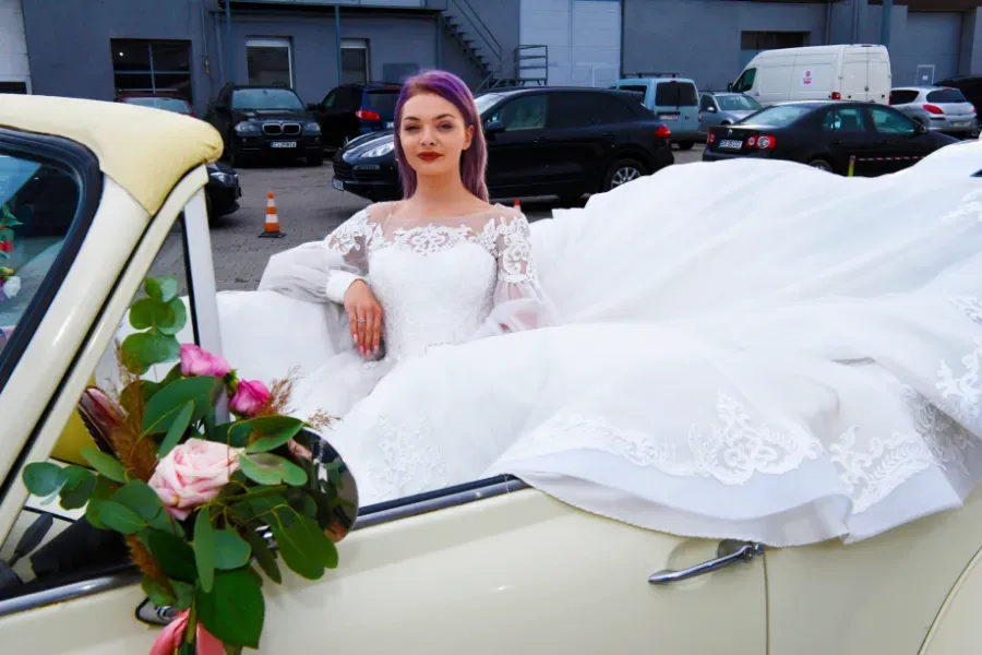 Novia con un vestido de novia de encaje blanco, asomada a un coche antiguo color crema con un ramo de flores, con un aparcamiento al fondo.
