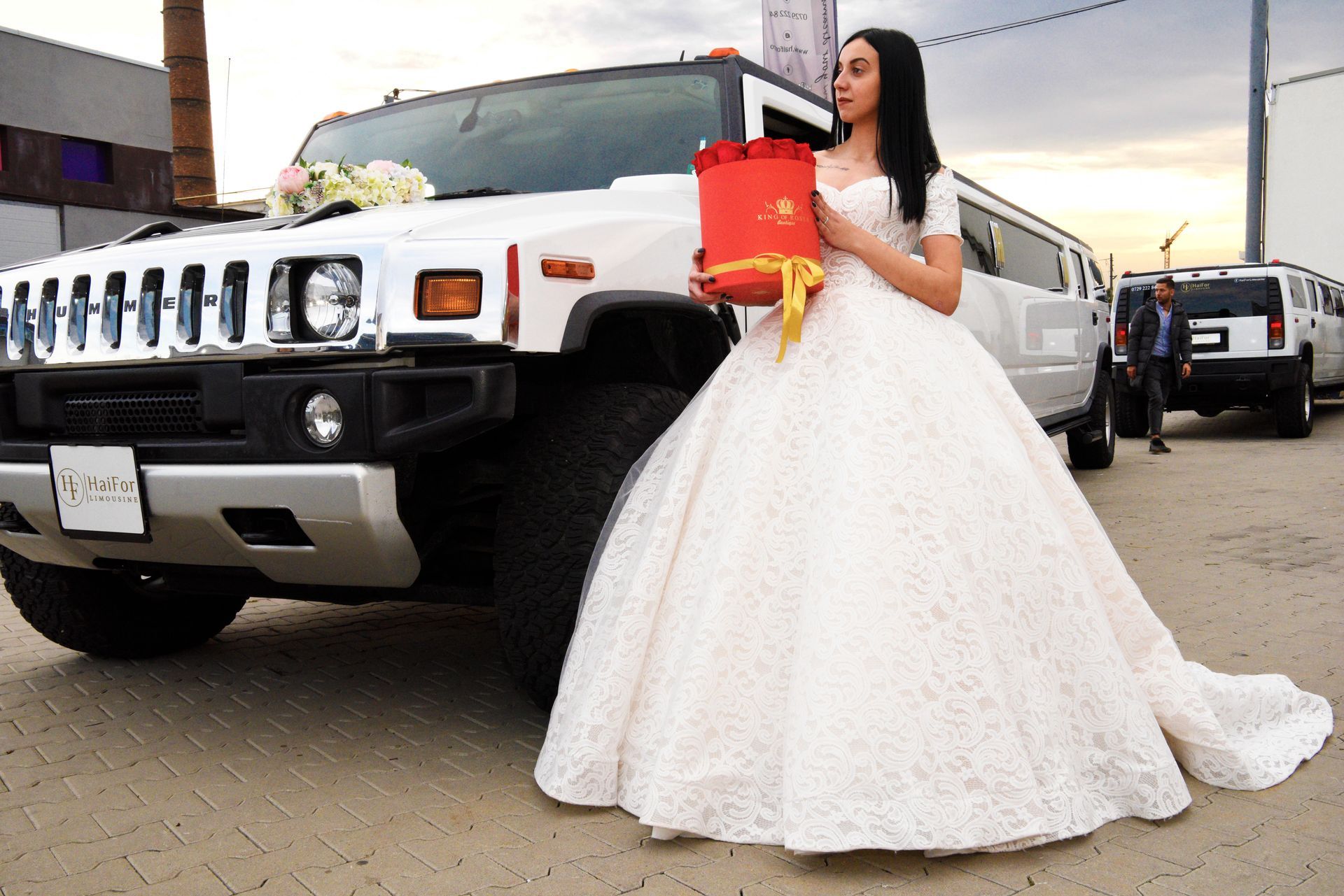 Mujer con un vestido de gala blanco sosteniendo una caja roja junto a una limusina Hummer blanca.
