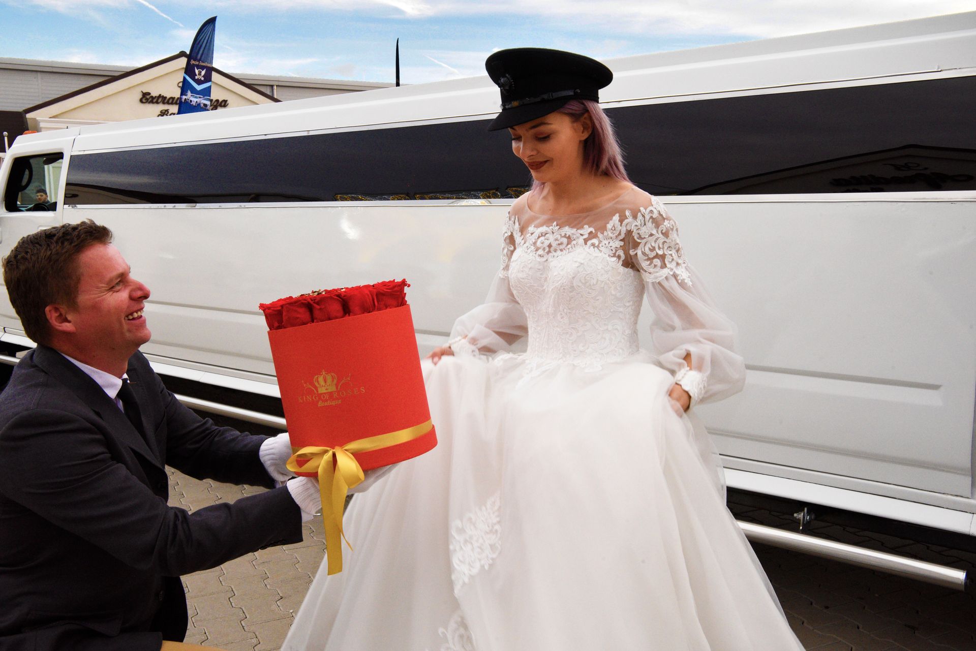Un hombre de traje le entrega una caja de regalo roja a una novia sonriente junto a una limusina blanca.