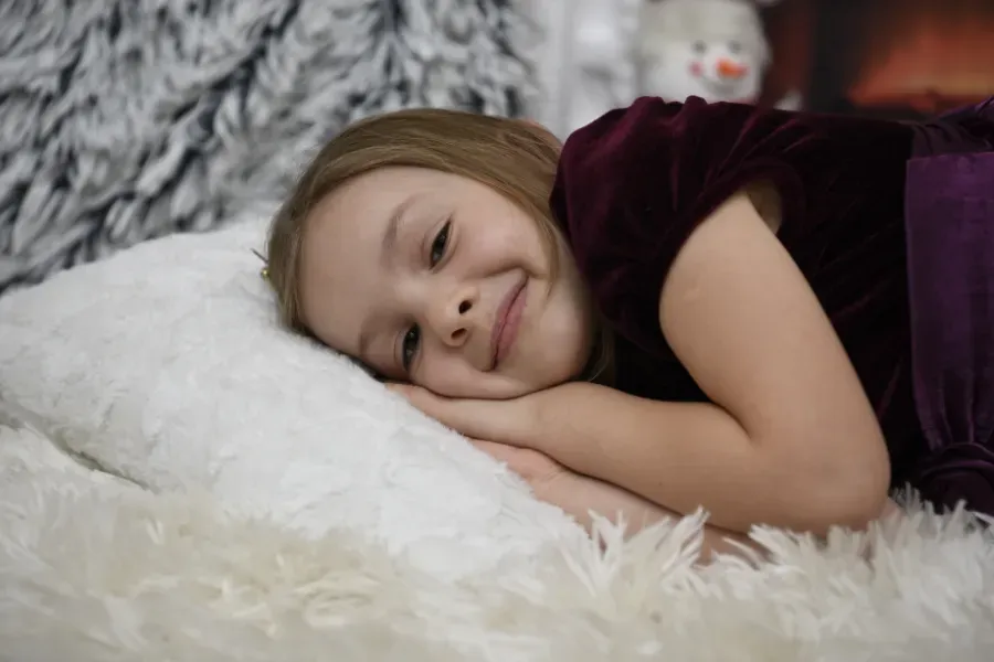Niña descansando sobre una suave manta blanca, sonriendo dulcemente con un vestido morado oscuro.