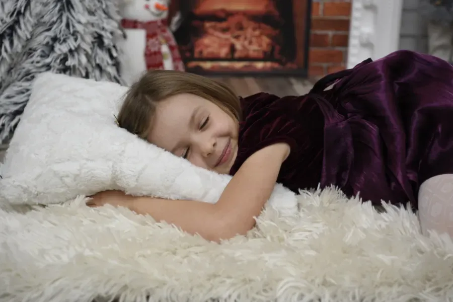 Un niño duerme en una cama blanca y mullida, apoyado sobre una almohada bajo una manta morada.