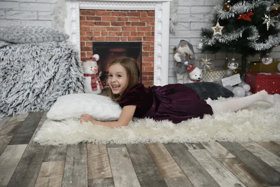 Una chica con un vestido color burdeos está tumbada sobre una alfombra junto a una chimenea y un árbol de Navidad, sonriendo a la cámara.