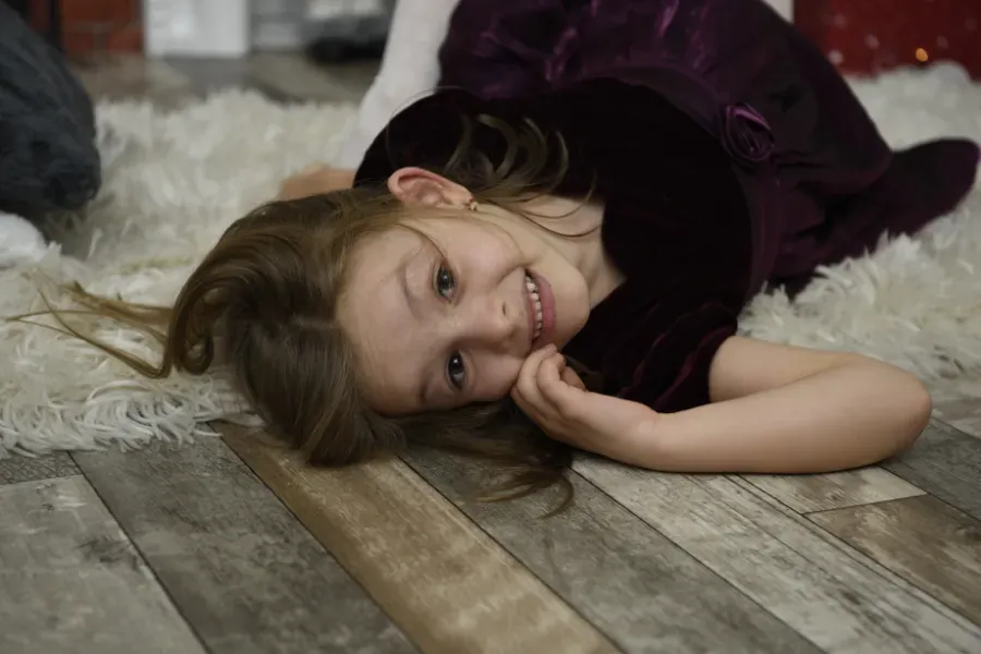 Niña sonriente tumbada sobre una alfombra junto a un suelo de madera, vestida con un vestido oscuro.