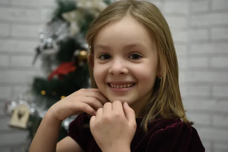 Niño sonriente con las manos cerca de la barbilla, frente a un árbol de Navidad borroso.