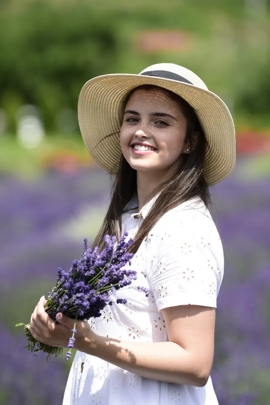 Mujer sonriente con sombrero de paja sosteniendo flores moradas en un campo de lavanda.