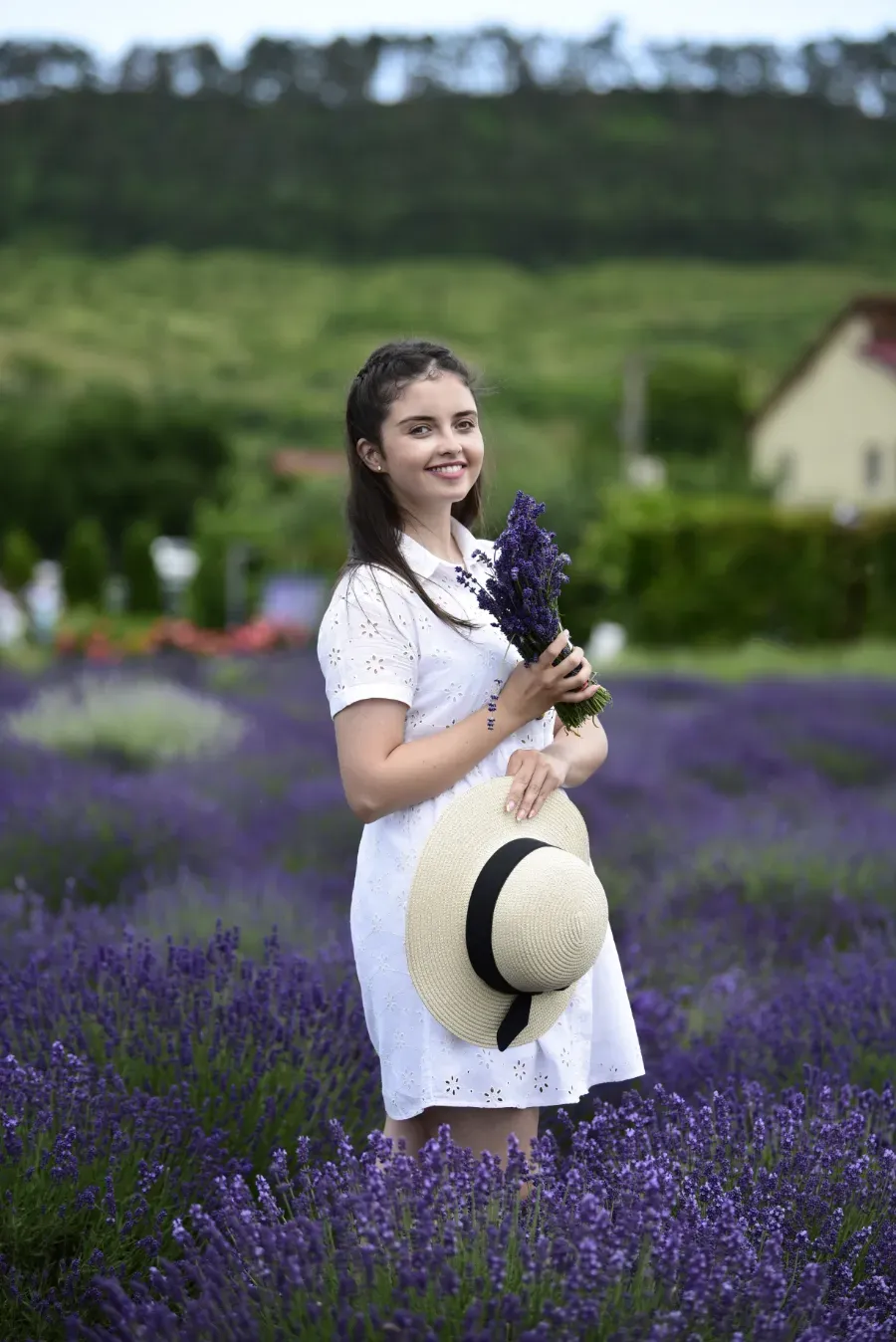 Mujer con vestido blanco de pie en un campo de lavanda morada, sosteniendo flores y un sombrero de paja.