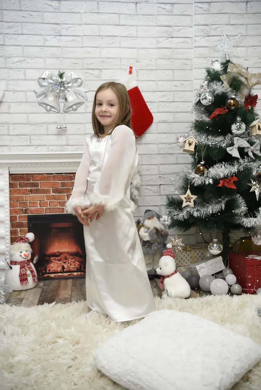 Una niña con un vestido blanco sonríe junto a una chimenea navideña y un árbol decorado, con un gorro de Papá Noel y cojines cerca.