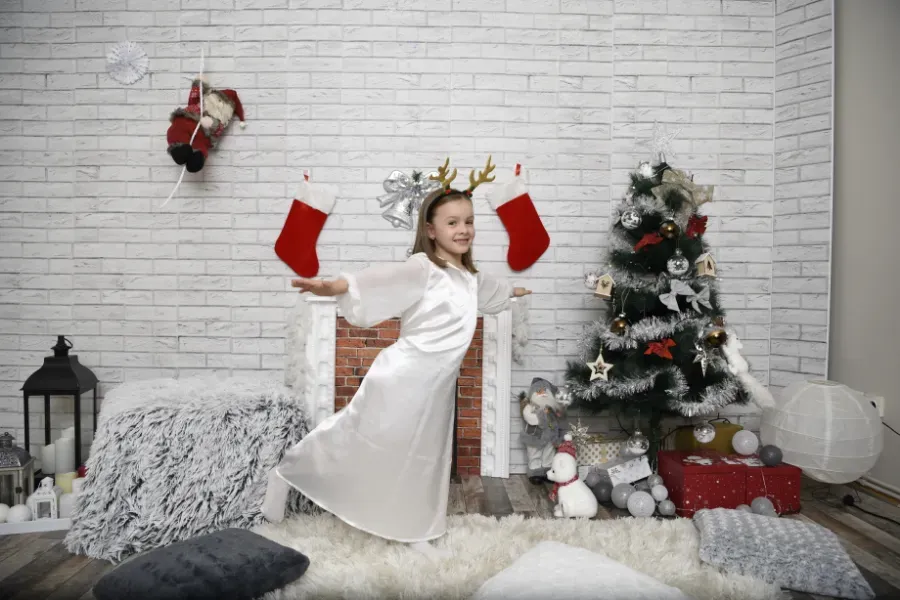 Una niña vestida de blanco con guantes rojos posa junto a un árbol de Navidad decorado y calcetines navideños en una acogedora sala de estar.