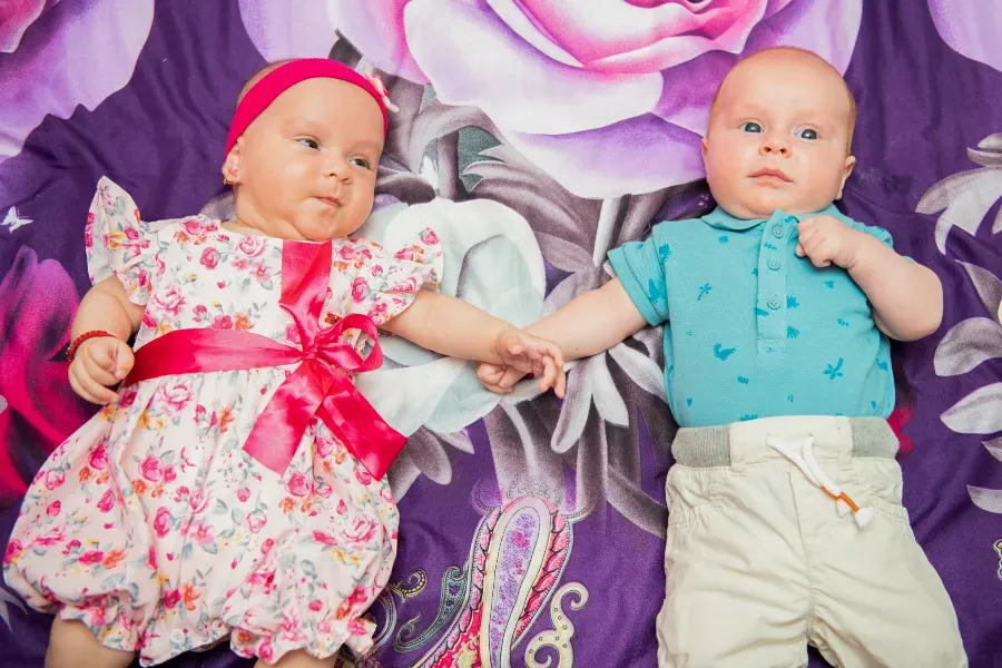 Dos bebés tomados de la mano sobre una manta estampada de flores; uno lleva un vestido rosa y el otro una camisa azul y pantalones beige.