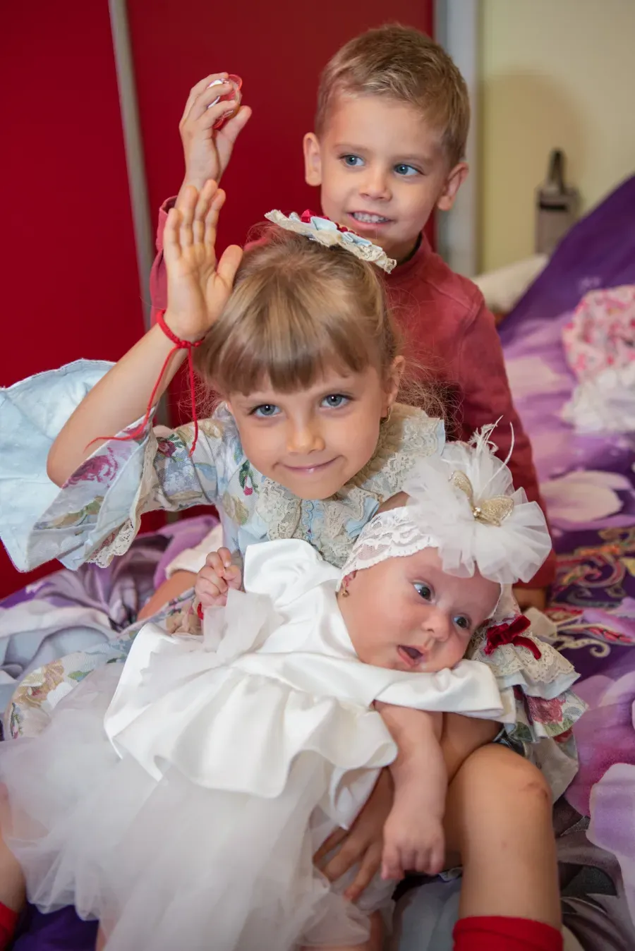 Tres niños vestidos con ropa blanca y burdeos posan juntos en una cama, uno de ellos sosteniendo una muñeca.