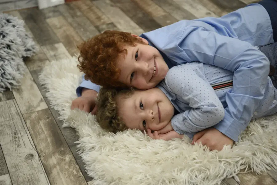 Dos niños tumbados sobre una alfombra mullida, uno con la cabeza apoyada en el hombro del otro y sonriendo.