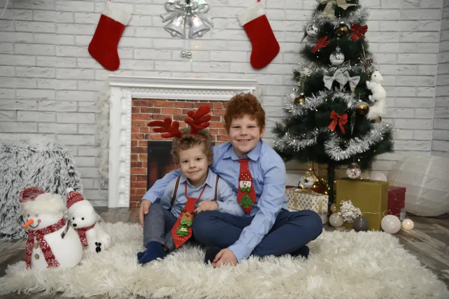 Dos niños sentados sobre una alfombra mullida junto a un árbol de Navidad decorado y calcetines navideños, frente a una chimenea.