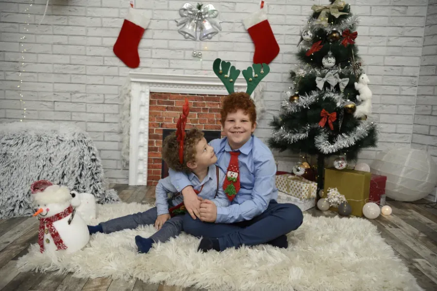 Un niño sentado sobre una alfombra mullida junto a un árbol de Navidad decorado, calcetines navideños y un muñeco de nieve en una habitación festiva.