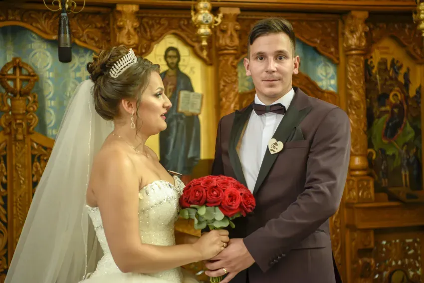 Los novios en una iglesia, sosteniendo un ramo rojo y mirando a la cámara.