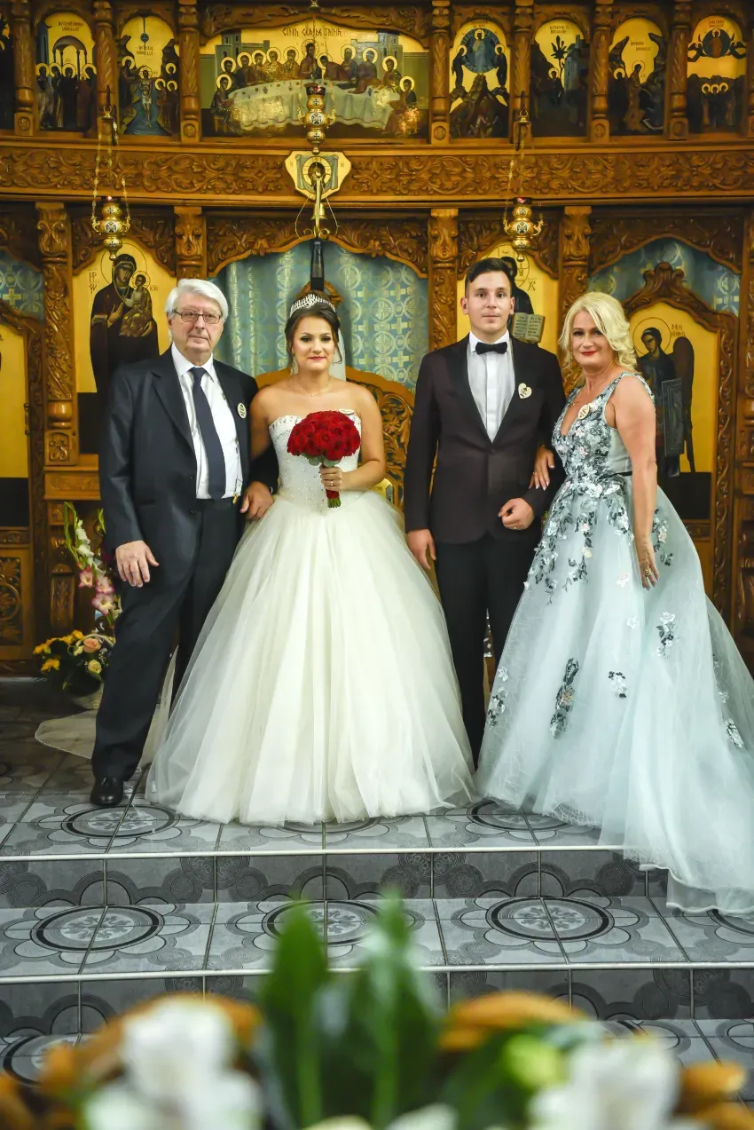 Cuatro personas posando con atuendo formal en una boda; la novia, vestida con un vestido blanco, sostiene flores rojas.