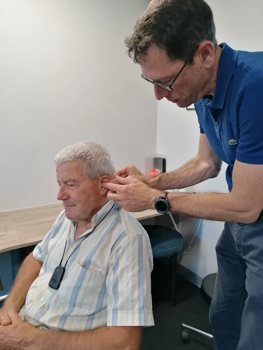 Un homme en polo bleu ajuste l’oreille d’un homme assis dans un cadre clinique