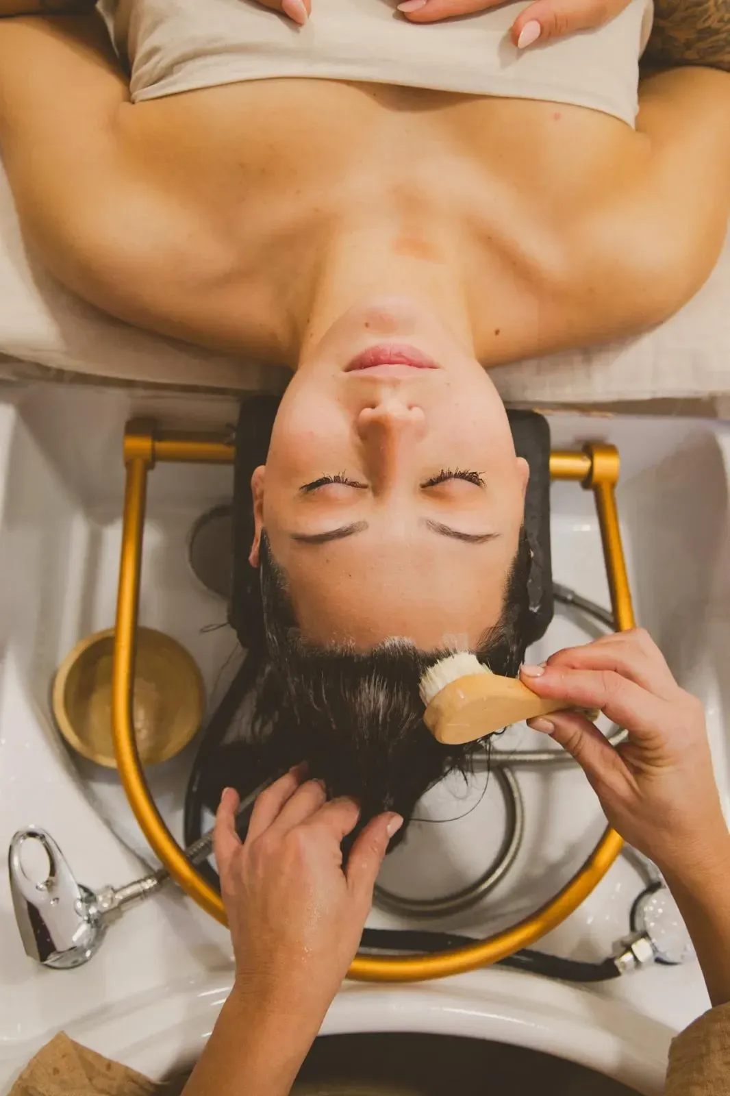 Mujer recibiendo un tratamiento capilar en un salón, con la cabeza apoyada en el lavabo y recibiendo un masaje con cepillo.