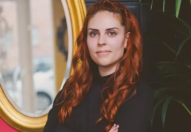Mujer con cabello rojo, brazos cruzados, parada frente a un espejo, top oscuro, entorno interior.