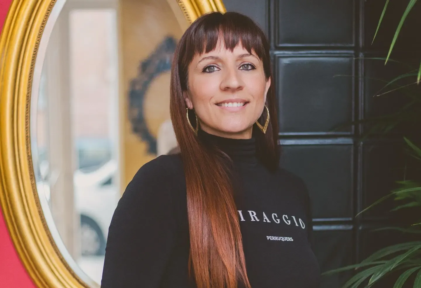 Mujer con cabello largo castaño y flequillo sonríe, vistiendo camisa negra y pendientes de oro, frente a un espejo.