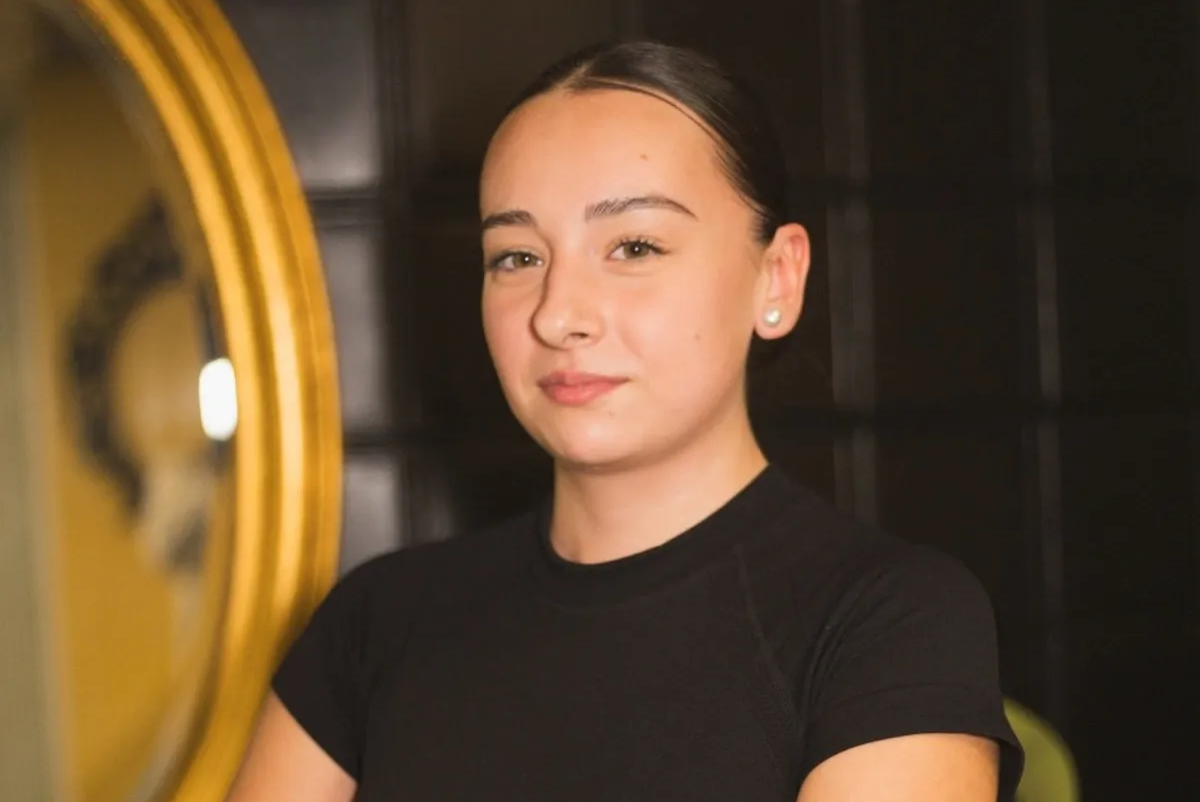 Mujer con top negro, cabello recogido, sonriendo levemente, cerca de un espejo con marco dorado.