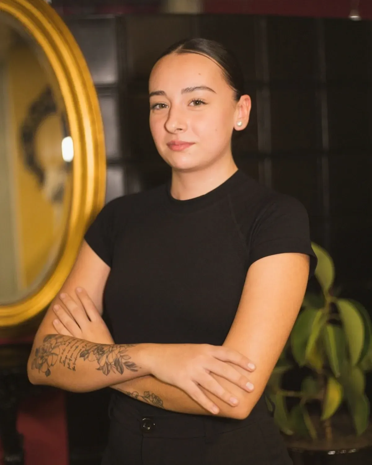 Mujer con los brazos cruzados, vestida con una camisa negra, parada frente a un espejo ovalado, tatuaje visible en el brazo.