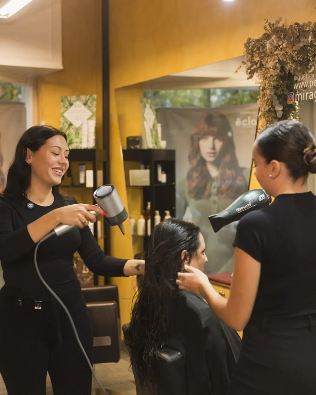 Dos estilistas secan el cabello mojado de una clienta en un salón. Una sonríe, la otra se concentra.