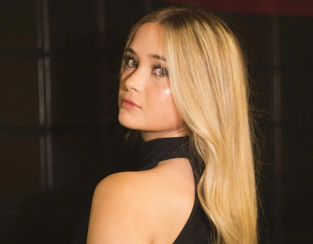 Mujer rubia de pelo largo mirando por encima del hombro. Fondo oscuro, vestida de negro.