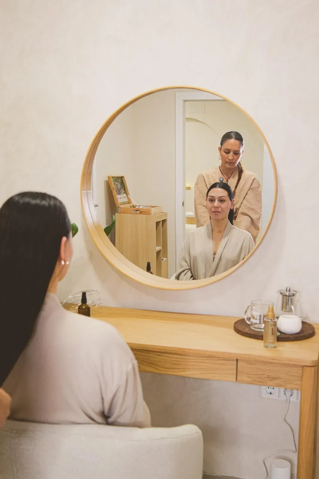 Una mujer en un salón de belleza se peina, reflejada en un espejo redondo. Otra persona la asiste desde atrás.