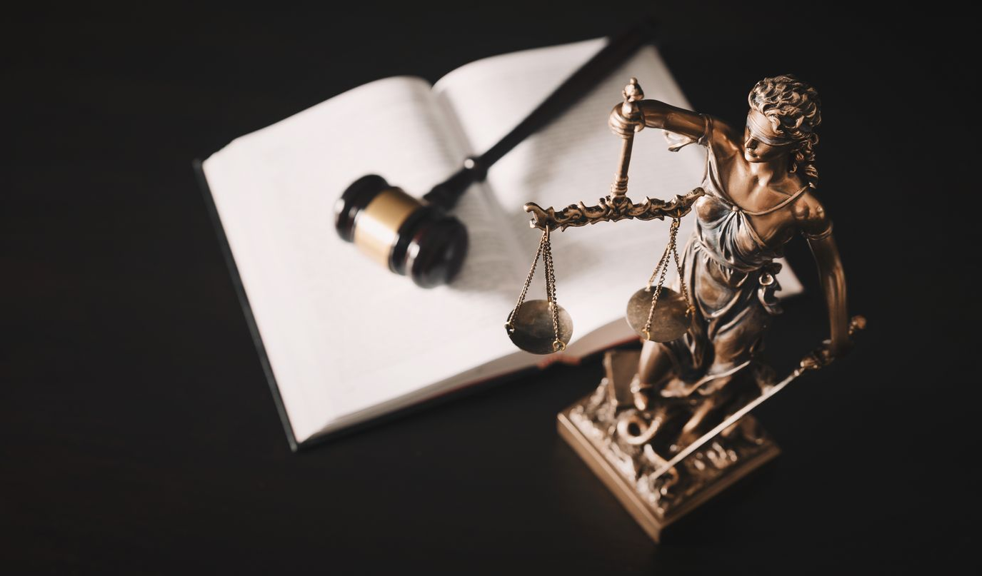 Statue de la Dame Justice avec balance, maillet et livre ouvert sur fond sombre.