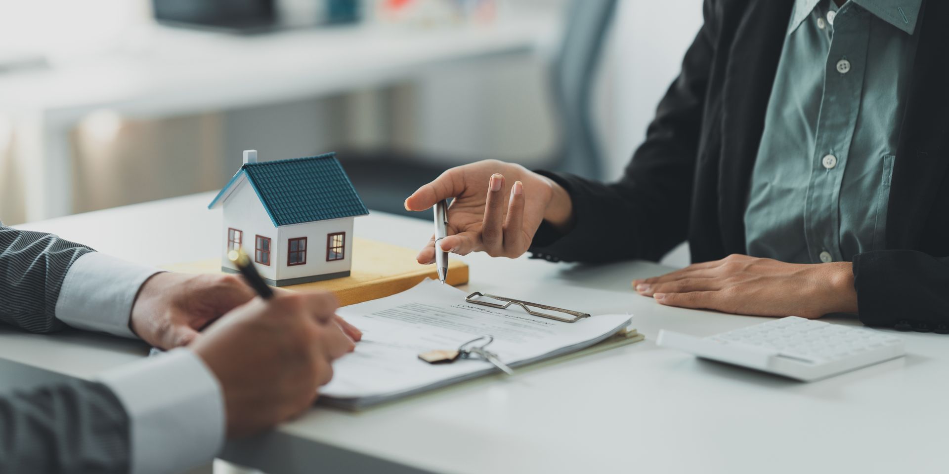 Deux personnes discutent d'une maquette de maison et de documents administratifs à un bureau lors d'une réunion immobilière.